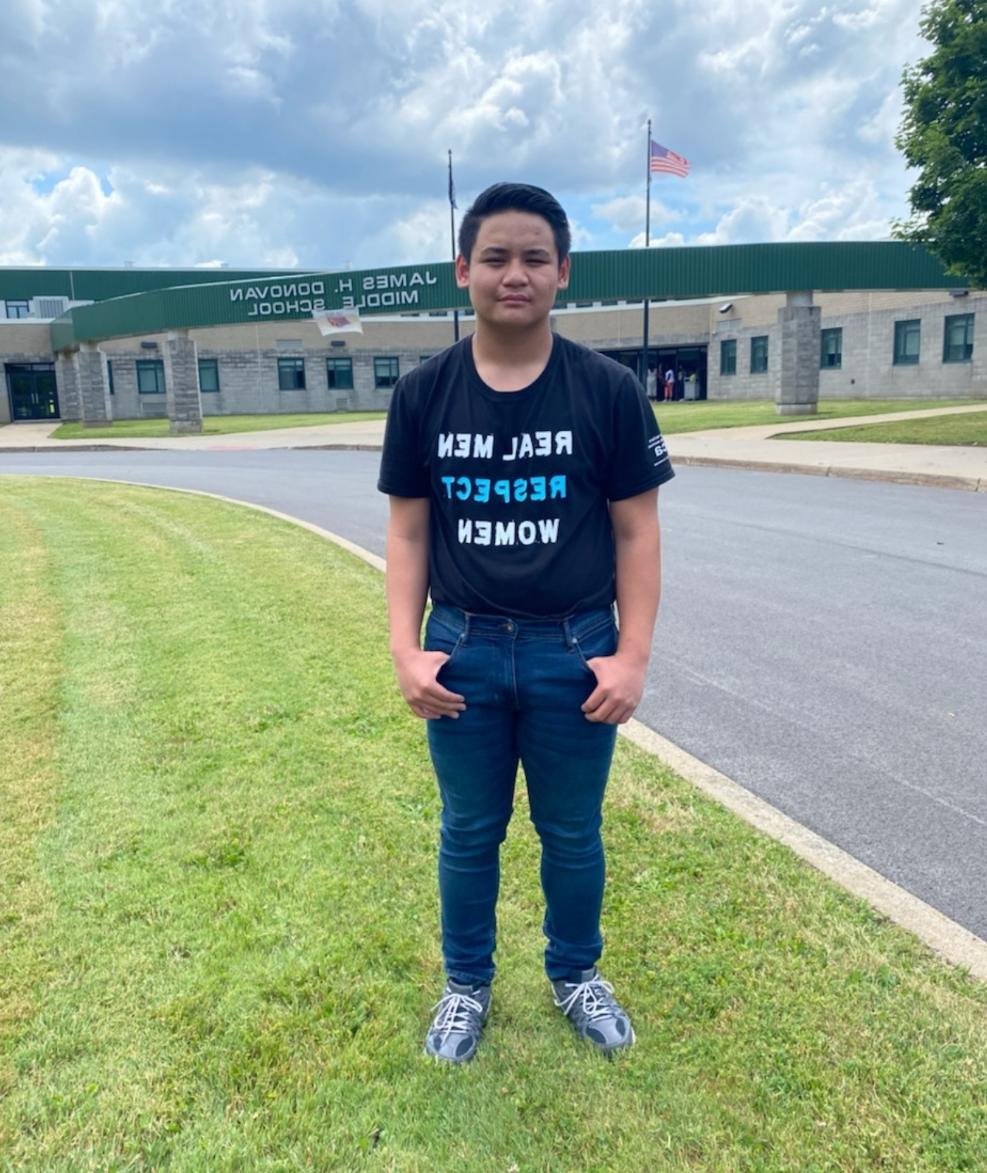 A young male student standing in front of Donovan Middle School wearing an AMEND Together shirt.