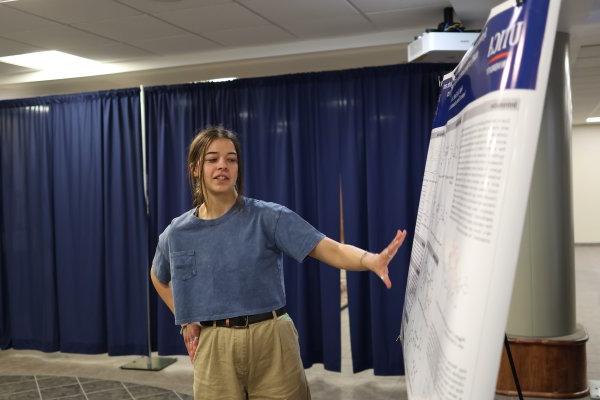 A student makes a presentation beside a poster at the 2024 Student Conference for Research, Professional Activities, and Creative Arts